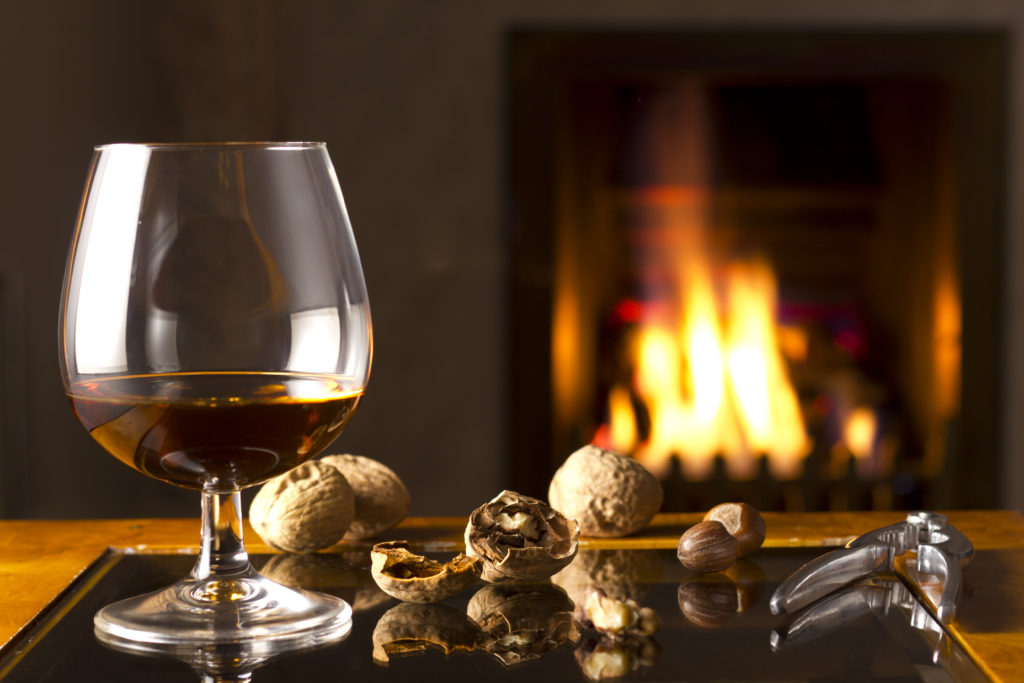 A Brandy snifter ready for drinking, placed next to cracked and uncracked hazelnuts with a stainless steel nutcracker by the fireplace.